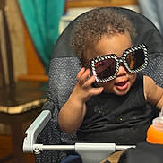 Xykie joined the competition — help win amazing prizes! toddler, child, sunglasses, high_chair, indoor, curtains, table, black_clothing, shorts, hand, face, hair, window, chair, furniture, playful, baby, person, glasses, smile