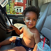 Daxaiireo is registered to the contest to win money with this photo: toddler, child, smiling, curly_hair, shirtless, shorts, car_interior, steering_wheel, seat, drink_cup, red_straw, sunlight, window, building, hand, face, portrait, casual, vehicle, happy