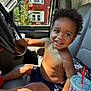 Daxaiireo is registered to the contest to win money with this photo: toddler, child, smiling, curly_hair, shirtless, vehicle_interior, steering_wheel, car_seat, drink_cup, sunlight, shadow, window, urban_background, red_brick_building, happy, casual, indoor, seatbelt, hand, face