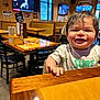 Xavier is registered to the contest to win money with this photo: toddler, child, smiling, indoor, restaurant, table, wooden_table, high_chair, casual_clothing, short_sleeves, hair, face, furniture, window, television, wall, light, seat, happy, person