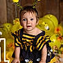 child, toddler, baby, bee_costume, headband, antennae, blue_eyes, smile, portrait, studio_photo, yellow_balloons, wooden_background, barrel, party_decoration, flowers, striped_dress, earrings, hands, cute, celebration