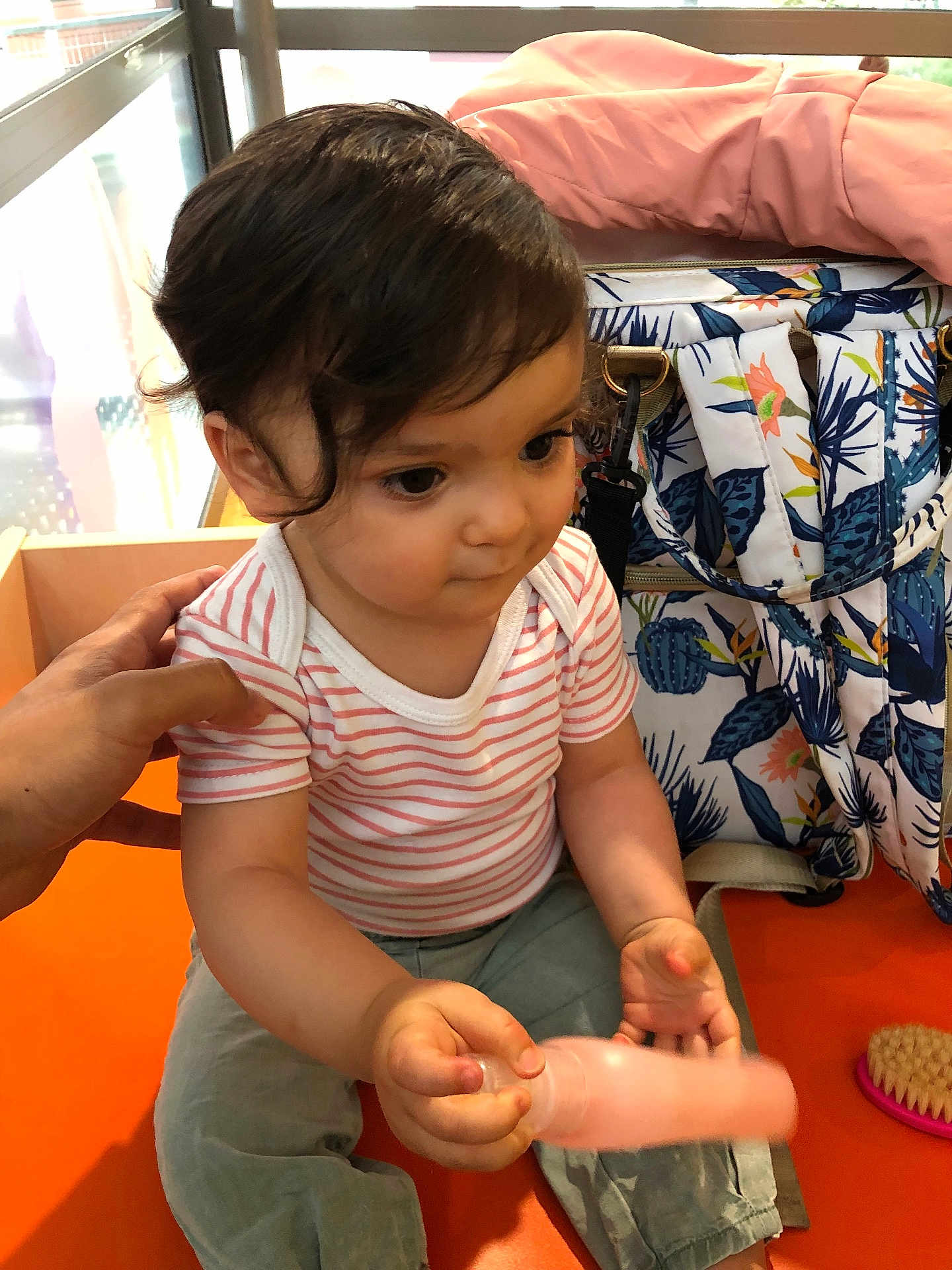 Nélia participe au concours pour gagner de l'argent avec cette photo : toddler, child, baby, bottle, hand, bag, brush, indoor, sitting, curly_hair, striped_shirt, pants, orange_surface, patterned_bag, person, cute, young_child, closeup, portrait, playful