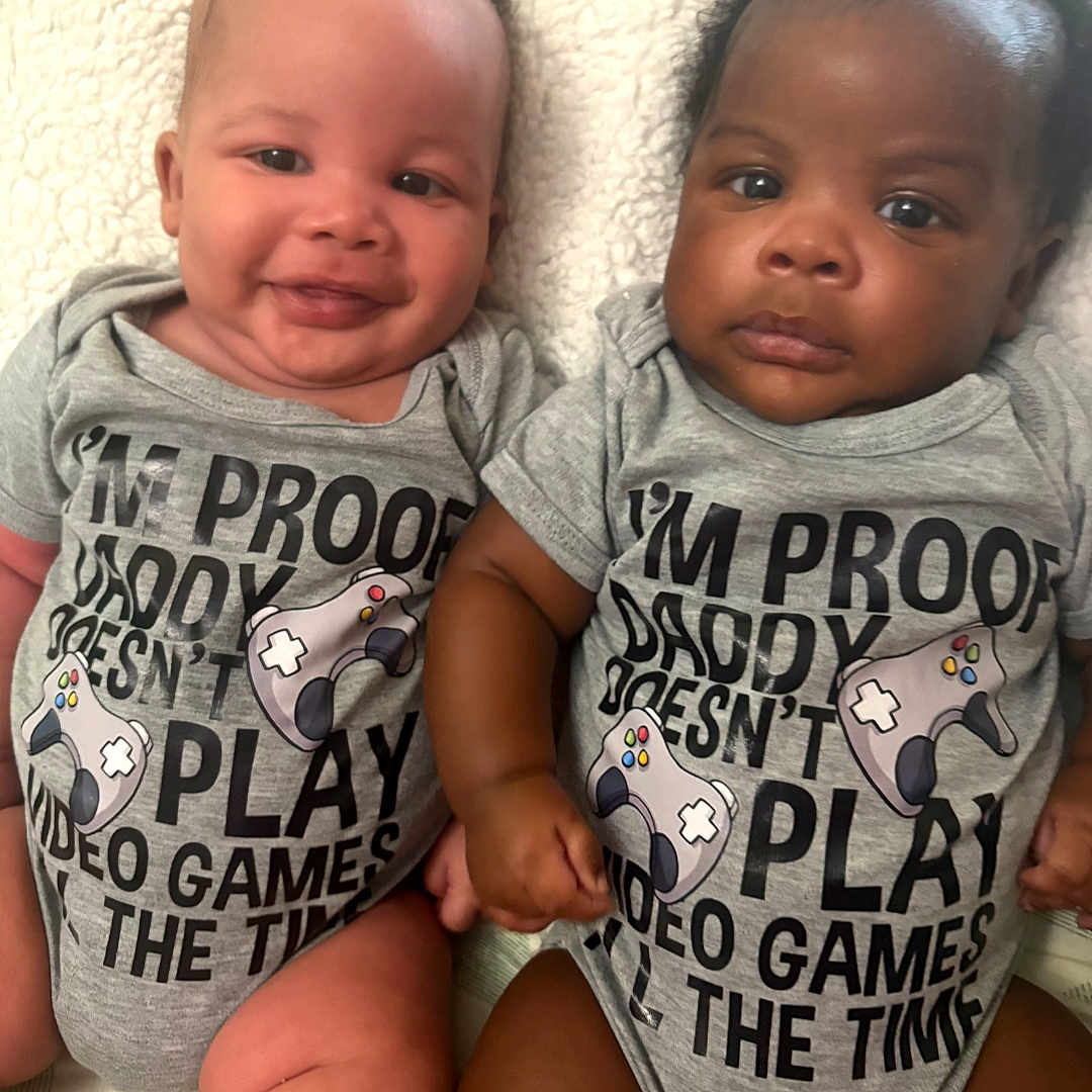 Keith And Keion joined the competition — help win amazing prizes! baby, blanket, closeup, clothing, controller, cute, diversity, gray, holding_hands, indoors, infant, onesie, portrait, serious, sitting, smiling, text, toddler, two_babies, video_games