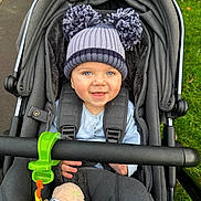Dante is registered to the contest to win money with this photo: baby, stroller, hat, pom_poms, smiling, child, blue_eyes, toy, plush_toy, green_clip, gray_fabric, outdoor, grass, pavement, cozy, winter_clothing, infant, cute, happy, portrait