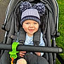 baby, stroller, hat, pom_poms, smiling, child, blue_eyes, toy, plush_toy, green_clip, gray_fabric, outdoor, grass, pavement, cozy, winter_clothing, infant, cute, happy, portrait