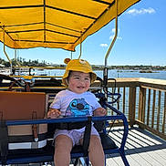 Sofia joined the competition — help win amazing prizes! baby, child, infant, smiling, outdoor, boat, pedal_boat, lake, water, dock, canopy, yellow_hat, bear_hat, sunglasses, stroller, safety_harness, seated, sunny, portrait, daylight