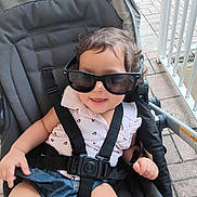 Alana Cobas joined the competition — help win amazing prizes! toddler, child, stroller, sunglasses, casual_clothing, denim_shorts, safety_harness, outdoor, pavement, railing, curly_hair, smiling, baby, seat, sunny, daytime, cute, relaxed, person, fashion