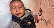 Zaylen is registered to the contest to win money with this photo: baby, bed, blanket, brown_hair, child, close_up, clothing, cozy, denim, face, happy, indoor, infant, lying_down, overalls, photography, portrait, smiling, soft_fabric, sweater