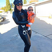 Nathan is registered to the contest to win money with this photo: baby, black_outfit, building, concrete, cool_pose, costume, daylight, fashion, footwear, handcuffs, holding, orange_clothing, outdoor, person, police_hat, shadow, sidewalk, sunglasses, trees, woman