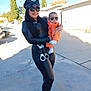 baby, black_outfit, building, concrete, cool_pose, costume, daylight, fashion, footwear, handcuffs, holding, orange_clothing, outdoor, person, police_hat, shadow, sidewalk, sunglasses, trees, woman