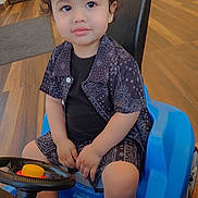 Nathan is registered to the contest to win money with this photo: child, toddler, toy_car, indoor, curly_hair, black_clothing, patterned_shirt, patterned_shorts, blue_toy, steering_wheel, flooring, wood_floor, sitting, young_child, cute, playful, small_vehicle, person, casual, expression