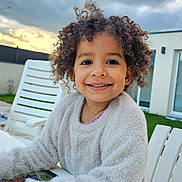 Zaiah a rejoint le concours — aidez-le/la à gagner de superbes lots ! child, smiling, curly_hair, sweater, white_table, outdoor, cloudy_sky, house, grass, chair, happy, person, portrait, young, casual_clothing, daytime, relaxed, nature, seating, playful