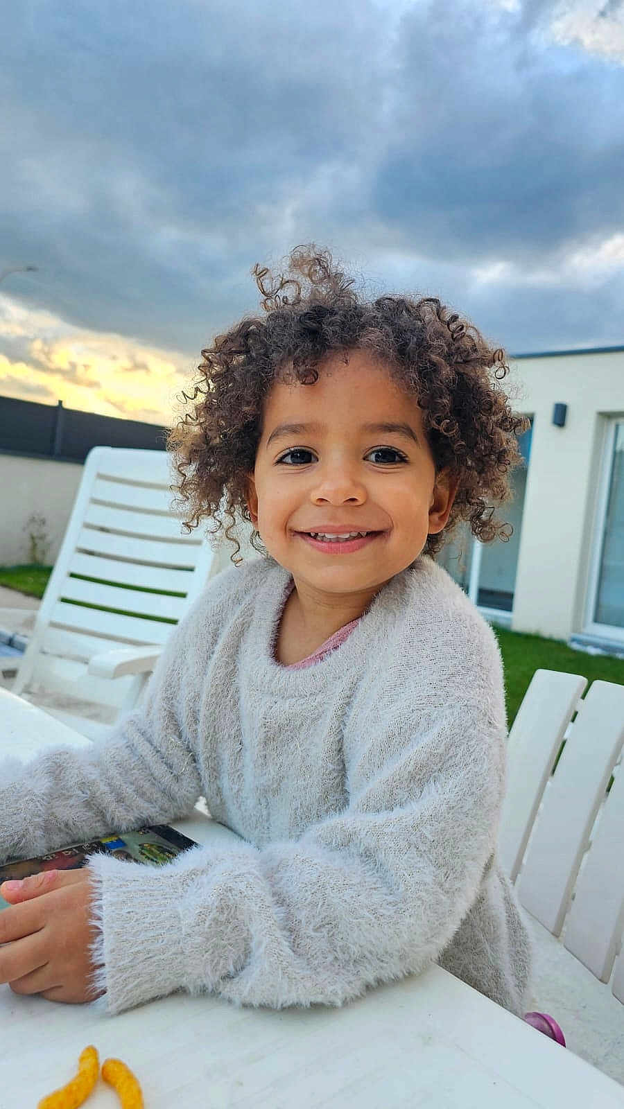 Zaiah a rejoint le concours — aidez-le/la à gagner de superbes lots ! child, smiling, curly_hair, sweater, white_table, outdoor, cloudy_sky, house, grass, chair, happy, person, portrait, young, casual_clothing, daytime, relaxed, nature, seating, playful