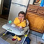 baby, child, walker, kitchen, refrigerator, wooden_cabinet, tile_floor, toy, snack, eating, yellow_jacket, socks, red_socks, seat, cute, portrait, indoor, appliance, backpack, slipper