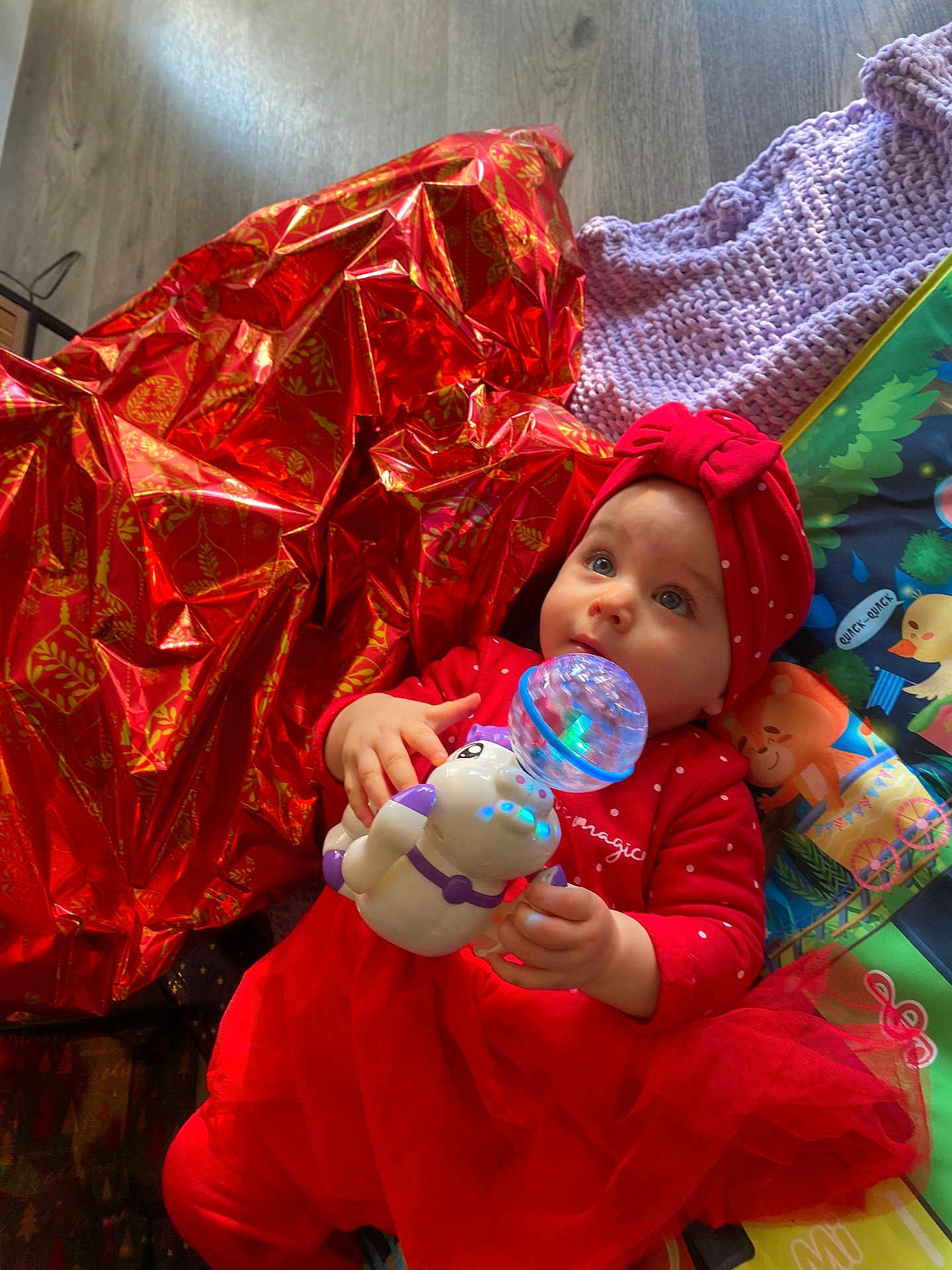 Sileyna participe au concours pour gagner de l'argent avec cette photo : baby, blue_eyes, child, colorful, curious, cute, face, festive, floor, glowing_toy, hands, headband, indoor, infant, knitted_blanket, lying_down, playmat, red_clothing, toy, wrapping_paper