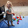 blue_mat, casual_clothing, chair, child, curly_hair, cute, daylight, fence, footwear, gravel, happy, outdoor, person, plastic_toys, play, playtime, smiling, standing, toys, young_child