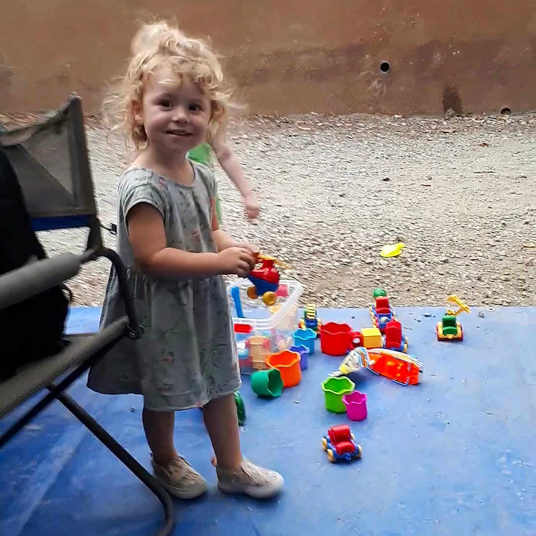 Lola participe au concours pour gagner de l'argent avec cette photo : blue_mat, casual_clothing, chair, child, curly_hair, cute, daylight, fence, footwear, gravel, happy, outdoor, person, plastic_toys, play, playtime, smiling, standing, toys, young_child