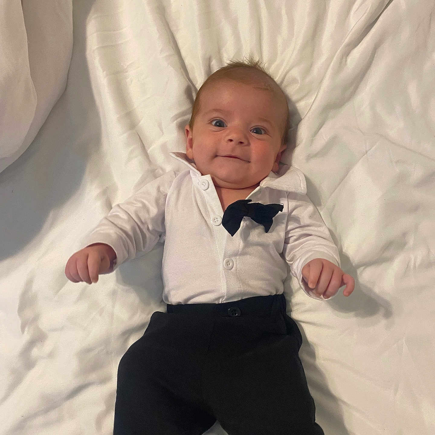Ashton is registered to the contest to win money with this photo: baby, infant, smile, white_shirt, black_pants, bow_tie, lying_down, blanket, cute, portrait, child, young, happy, indoors, clothing, person, face, small_hands, looking_at_camera, tuxedo