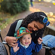 Everett is registered to the contest to win money with this photo: baby, infant, adult, parent, smiling, happy, portrait, outdoor, park, stroller, hat, sunglasses, curly_hair, blue_jacket, colorful_clothing, family, caregiver, child, nature, bokeh