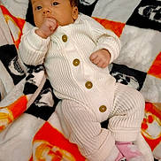 Riley joined the competition — help win amazing prizes! baby, infant, knitted_clothing, white_onesie, brown_buttons, pink_socks, skull_pattern, orange_blanket, black_blanket, white_blanket, lying_down, hand_in_mouth, curious_expression, soft_texture, cozy, indoor, child, cute, warm, portrait