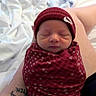 newborn, baby, sleeping, swaddle, beanie, red_clothing, tattoo, arm, lap, blanket, bed, indoor, portrait, person, infant, resting, peaceful, cute, closeup, human