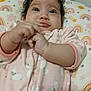 baby, infant, pink_onesie, sheep_pattern, cushion, rainbow_pattern, star_pattern, hands, face, eyes, curious, lying_down, indoors, child, cute, soft_toys, sleepwear, portrait, chubby_cheeks, cozy