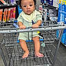baby, shopping_cart, store_aisle, water_dispenser, household_items, curly_hair, infant, green_outfit, bib, barefoot, metal_cart, floor, product_shelves, retail, child, young_child, indoor, shopping, holding, serious_expression