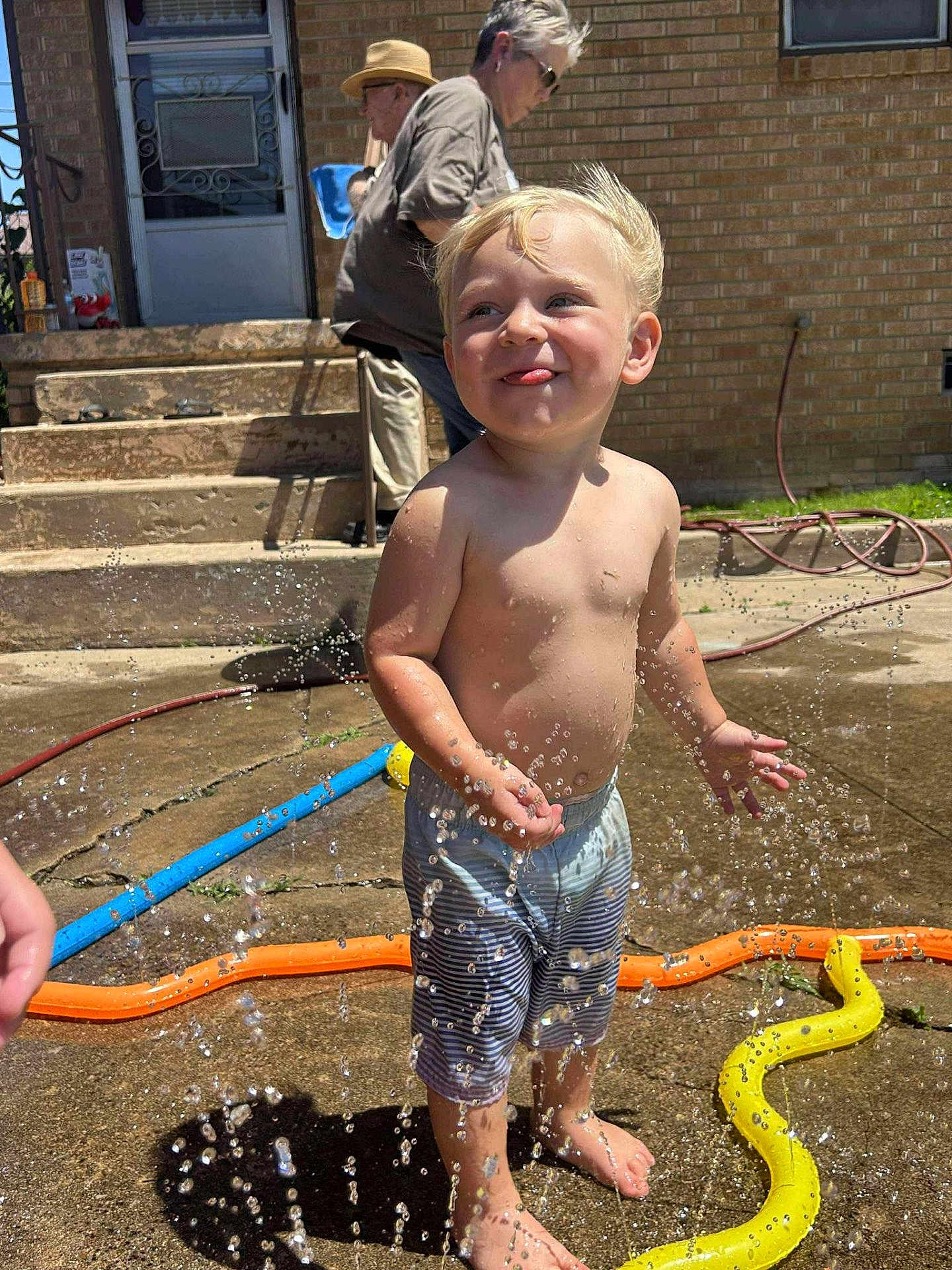 Cameron joined the competition — help win amazing prizes! toddler, child, water, sprinkler, summer, barefoot, play, outdoor, sunlight, brick_wall, concrete, hose, water_droplets, happy, smiling, shorts, people, adult, family, backyard