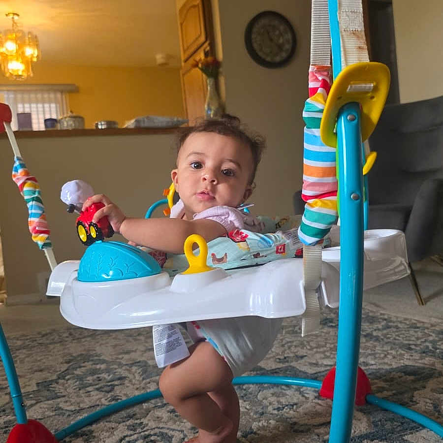 Esmail is registered to the contest to win money with this photo: baby, infant, child, face, person, sitting, activity_center, jumper, baby_toy, toy_car, colorful, indoor, living_room, carpet, furniture, chair, home, portrait, curious, playtime