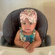 Sophia is registered to the contest to win money with this photo: baby, child, high_chair, bow, headwear, floral_pattern, hands, expression, face, indoor, clothing, sitting, cute, infant, person, portrait, furniture, table, skin, young