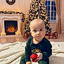 baby, christmas_tree, christmas_ornament, fireplace, wreath, gift_box, blanket, indoor, holiday, festive, decorations, pajamas, child, smiling, holiday_lights, curtains, floor, window, cozy, winter