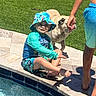 child, pool, water, swimwear, hat, dog, grass, sidewalk, sunny, outdoor, happy, smile, barefoot, person, summer, playing, pet, blue, sitting, fun