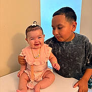 Valeria is registered to the contest to win money with this photo: baby, child, smiling, boy, sibling, indoor, table, necklace, pacifier, hair_tie, clothing, cute, happy, portrait, people, person, skin, face, hand, tabletop