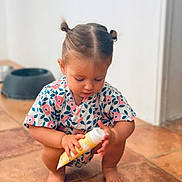 Adèle participe au concours pour gagner de l'argent avec cette photo : toddler, child, baby, squatting, floor, tile_floor, hair_buns, floral_clothing, indoor, skin, barefoot, toy, container, cream_tube, focus, curious, person, young_child, pet_bowl, background_blur