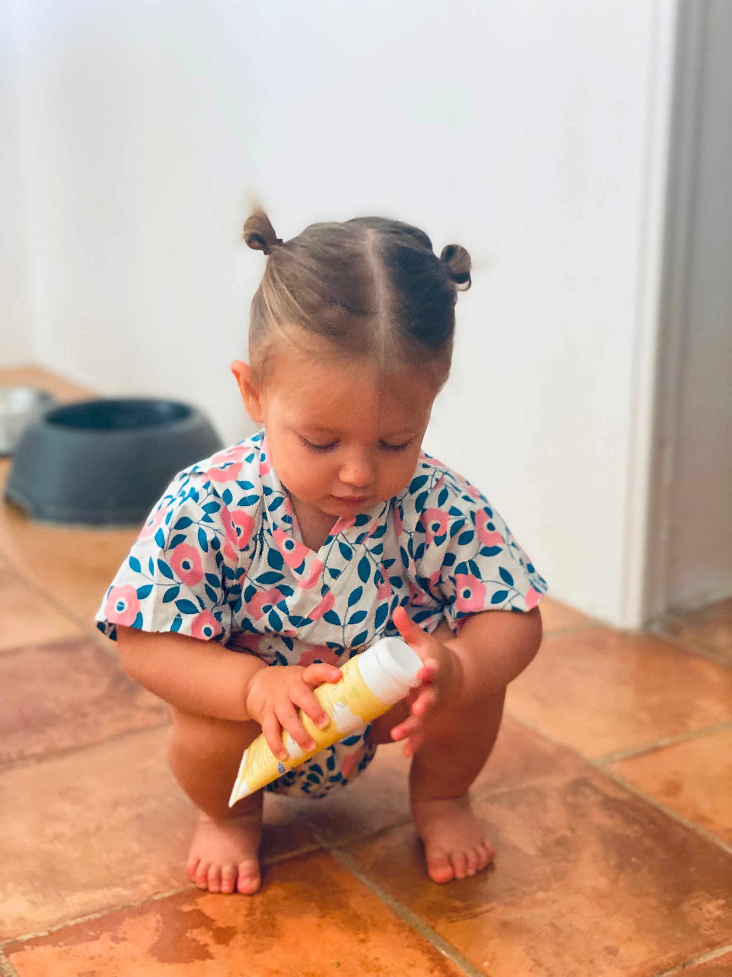 Adèle participe au concours pour gagner de l'argent avec cette photo : toddler, child, baby, squatting, floor, tile_floor, hair_buns, floral_clothing, indoor, skin, barefoot, toy, container, cream_tube, focus, curious, person, young_child, pet_bowl, background_blur