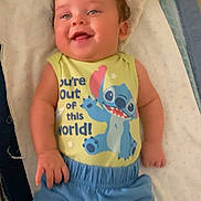 Abel is registered to the contest to win money with this photo: baby, child, smiling, happy, curly_hair, cartoon_character, clothing, blue_shorts, yellow_shirt, text_on_clothing, blanket, lying_down, indoor, cute, infant, person, portrait, soft_lighting, toddler, cozy