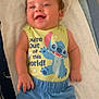 baby, child, smiling, happy, curly_hair, cartoon_character, clothing, blue_shorts, yellow_shirt, text_on_clothing, blanket, lying_down, indoor, cute, infant, person, portrait, soft_lighting, toddler, cozy