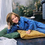 Luka participe au concours pour gagner de l'argent avec cette photo : child, smiling, denim_jacket, blue_jeans, cushions, green_cushion, yellow_cushion, indoor, curtains, floral_background, happy, casual_clothing, portrait, relaxed_pose, floor, wooden_floor, furniture, home_decor, person, young