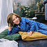 child, smiling, denim_jacket, blue_jeans, cushions, green_cushion, yellow_cushion, indoor, curtains, floral_background, happy, casual_clothing, portrait, relaxed_pose, floor, wooden_floor, furniture, home_decor, person, young