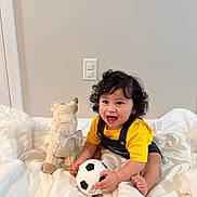 Gabriel joined the competition — help win amazing prizes! child, toddler, baby, smiling, happy, curly_hair, portrait, sitting, bed, bedding, soccer_ball, plush_toy, llama, yellow_shirt, overalls, indoors, bedroom, wall, light_switch, playtime