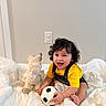 child, toddler, baby, smiling, happy, curly_hair, portrait, sitting, bed, bedding, soccer_ball, plush_toy, llama, yellow_shirt, overalls, indoors, bedroom, wall, light_switch, playtime
