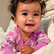 Ivy is registered to the contest to win money with this photo: baby, child, curly_hair, smiling, pink_clothing, onesie, bed, blanket, happy, cute, portrait, infant, indoors, face, person, young_child, resting, hand, eyelashes, cozy