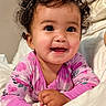 baby, child, curly_hair, smiling, pink_clothing, onesie, bed, blanket, happy, cute, portrait, infant, indoors, face, person, young_child, resting, hand, eyelashes, cozy