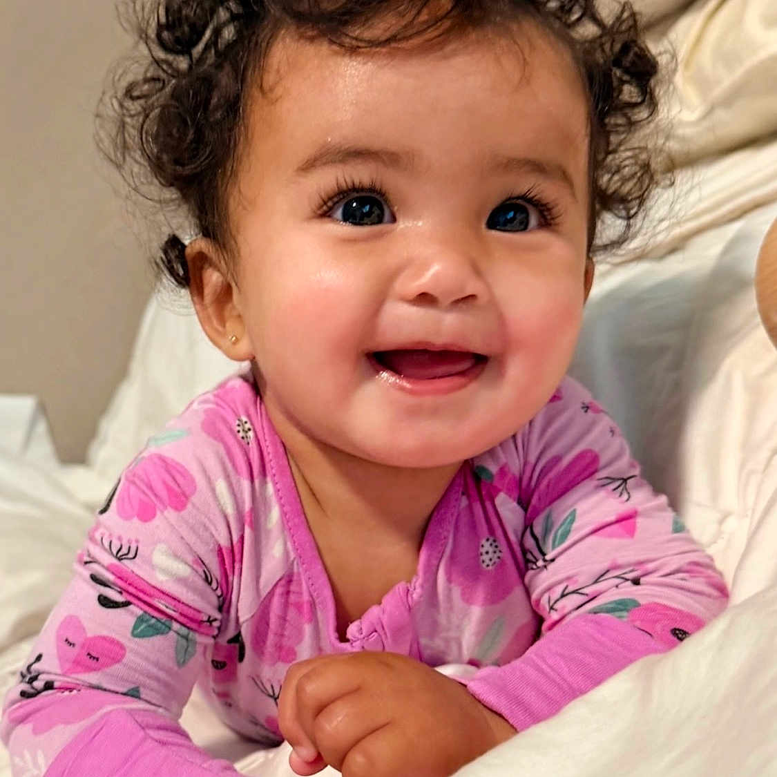 Ivy is registered to the contest to win money with this photo: baby, child, curly_hair, smiling, pink_clothing, onesie, bed, blanket, happy, cute, portrait, infant, indoors, face, person, young_child, resting, hand, eyelashes, cozy