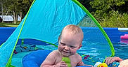 Evelyn joined the competition — help win amazing prizes! baby, child, pool, water, float, inflatable, toy, smile, outdoor, summer, blue, green, sunshade, playful, happy, person, nature, trees, sky, hand