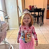 child, girl, toddler, princess, tiara, smiling, portrait, indoors, home, pink_clothing, dress, cartoon_character, necklace, bracelet, red_hair, standing, baby_bouncer, dining_table, tile_floor, sunlight