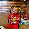 child, toddler, girl, smiling, red_hair, hair_bows, grinch, christmas, sweatshirt, red_clothing, candy_cane, sitting, chair, indoor, wood_paneling, carpet, television, beverages, water_bottles, portrait