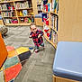 toddler, child, library, bookshelf, books, plaid_shirt, carpet, colorful, indoor, walking, mismatched_socks, furniture, reading_area, young_child, person, floor, seat, plush_toy, educational, playful