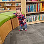 toddler, child, library, bookshelf, books, plaid_shirt, socks, carpet, cushioned_seat, wooden_furniture, indoor, person, floor, young_child, walking, cute, colorful, education, learning, room