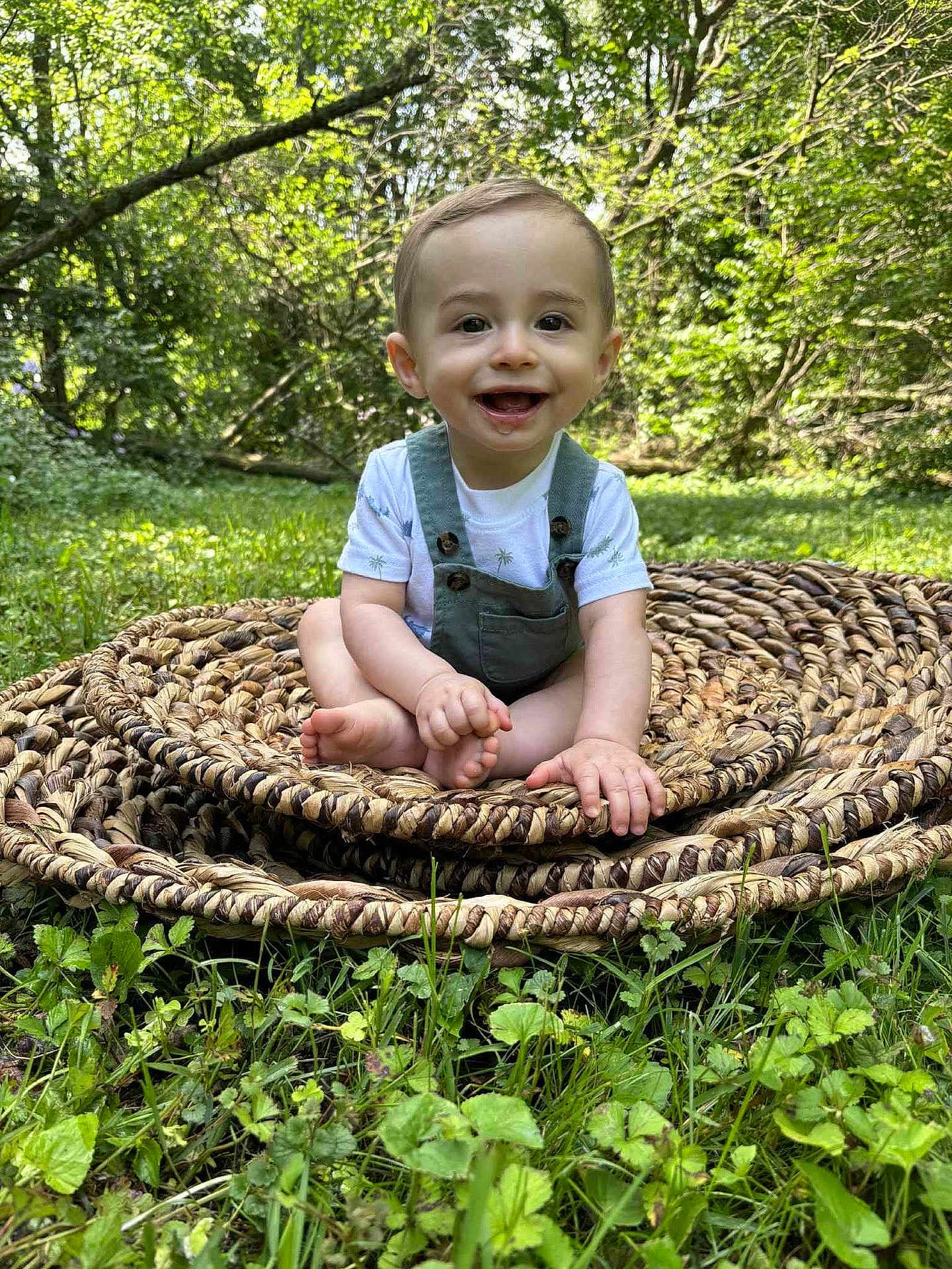 Robbie is registered to the contest to win money with this photo: baby, child, smile, outdoor, grass, greenery, nature, woven_mat, sitting, happy, portrait, infant, cute, sunlight, trees, young, person, summer, playful, casual_clothing